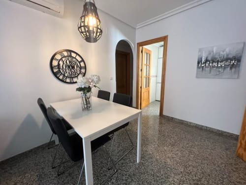 Comedor moderno en apartamento de Fuengirola con mesa blanca, sillas negras, reloj decorativo y lámpara colgante