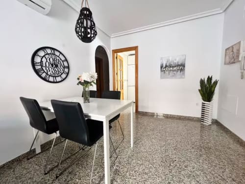 Comedor moderno de apartamento en Fuengirola con mesa blanca, sillas negras, reloj de pared geométrico y decoración contemporánea