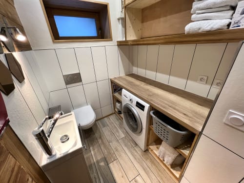 Modern laundry room with washing machine, dryer, white subway tile walls, wood shelving with towels, and small sink at Chez