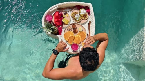 Aerial view of person in turquoise water holding breakfast tray with fruits, pastries and flowers at Oasis Dune Villas Lombok