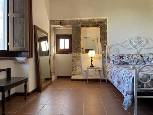 Spacious bedroom at Mater Familias Oasis in Siracusa with ornate white bed, rustic stone archway, and terracotta tile floor.