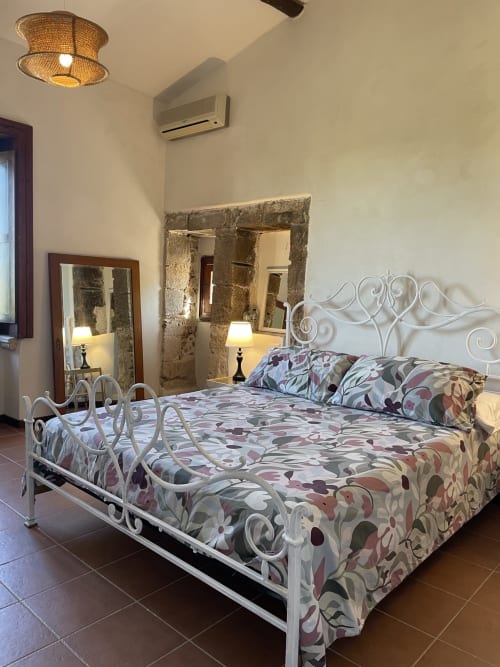 Camera elegante presso Mater Familias Oasis a Siracusa con letto in ferro battuto bianco ornamentale, biancheria floreale e specchio con cornice in pietra