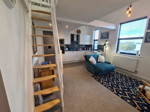 Open-plan living space at 2 Doves Holiday Property, Exeter, with kitchen, blue chaise lounge, and wooden loft stairs.