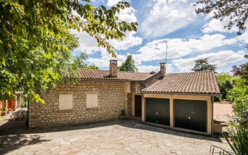 Villa en pierre Casa de la huerta à Millau, toit en terre cuite, garages et allée, entourée de verdure sous un ciel dégagé