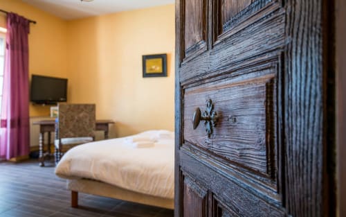 Porte rustique en bois ouvrant sur une chambre moderne avec lit blanc, murs jaunes, télévision et rideaux roses à la villa de Millau