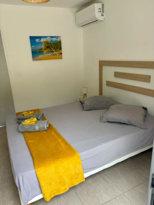 Chambre lumineuse à Maly Blue Location au Moule avec lit queen, serviettes jaunes, coussins gris, climatisation murale et ambiance tropicale