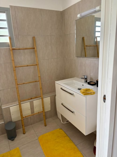 Salle de bains moderne avec vanité blanche, murs en carrelage beige, échelle en bambou et accents jaunes à Maly Blue, Guadeloupe