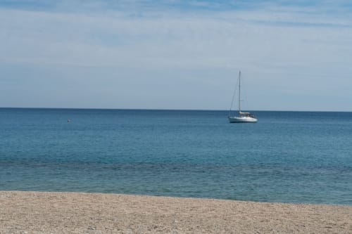 Barca a vela ancorata al largo della spiaggia di Varigotti con mare turchese calmo e riva sabbiosa, vista dalla camera Ancora