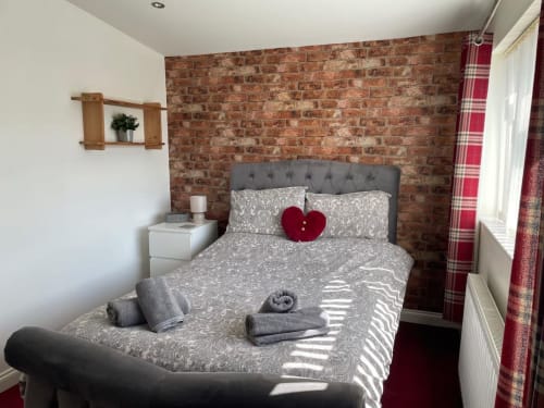 Cozy bedroom at 2 Doves Holiday Property in Exeter with brick accent wall, gray upholstered bed, and red plaid curtains