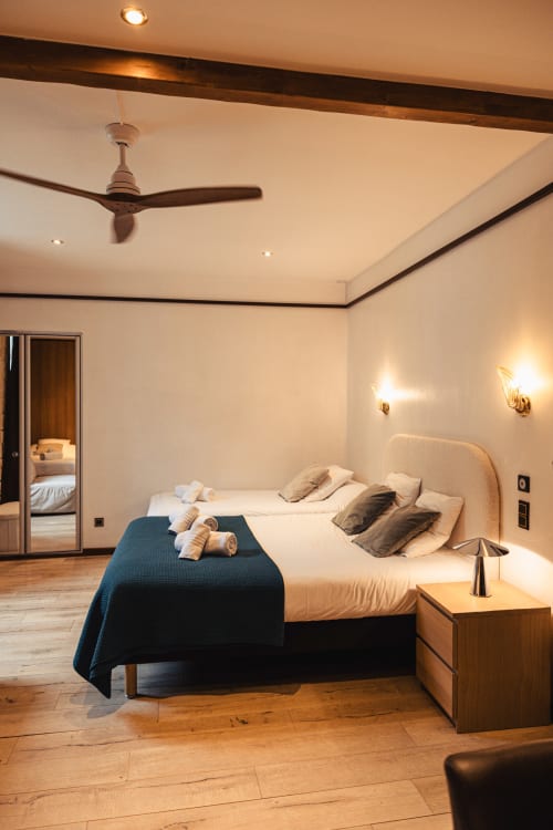 Spacieuse chambre familiale du Savoy Hôtel avec deux lits, table de nuit en bois, ventilateur au plafond et appliques murales au Bourget-du-Lac