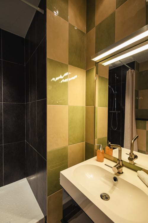 Salle de bains moderne de l'Hôtel Savoy avec carrelage olive et crème, lavabo blanc et paroi de douche noire