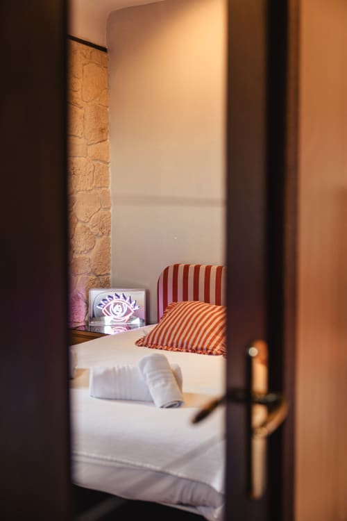 Chambre du Savoy Hôtel avec coussins rayés, mur en pierre et œuvre d'art néon représentant un œil visible par la porte
