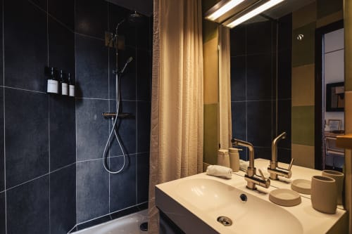 Salle de bain moderne du Savoy Hôtel avec douche carrelée foncée, double vasque, robinetterie en laiton et éclairage chaleureux au