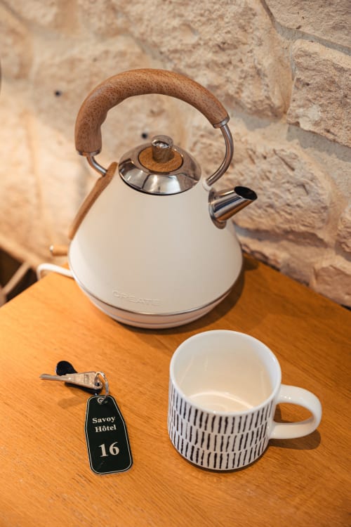 Équipements chambre Savoy Hôtel : bouilloire blanche à poignée bois, tasse à motifs et clé de chambre sur surface bois