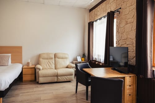Spacieuse chambre d'hôtel au Savoy Hôtel avec canapé crème, bureau en bois avec TV, lit et mur en pierre à Le Bourget-du-Lac