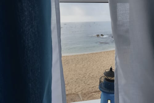 Vue sur la mer depuis le balcon d'un appartement en bord de plage à Palafrugell, avec plage de sable et eaux méditerranéennes visibles à travers les rideaux