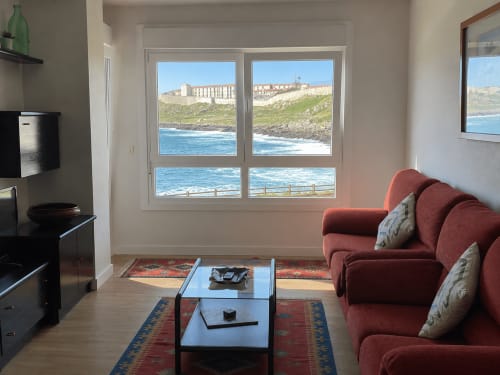 Moderno salón con vistas al mar de la costa de Baiona a través de grandes ventanales, sofá burdeos y mobiliario contemporáneo.