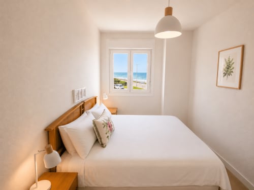 Dormitorio costero luminoso con vistas al mar, cama de madera, lámpara colgante y playa de Baiona visible desde las ventanas