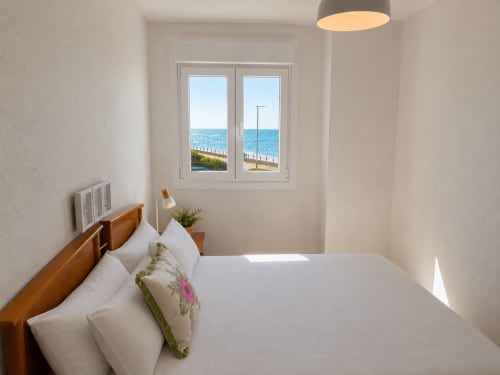 Luminoso dormitorio costero en Baiona con vistas al mar a través de ventanales dobles, cama de madera con sábanas blancas e iluminación de techo moderna