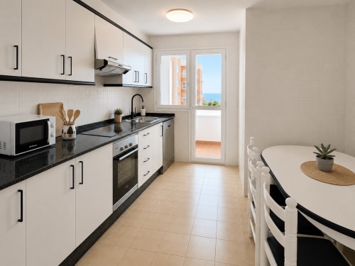 Cocina moderna con encimeras de granito negro, armarios blancos, mesa de comedor y puertas con vistas al mar en el apartamento As Estelas