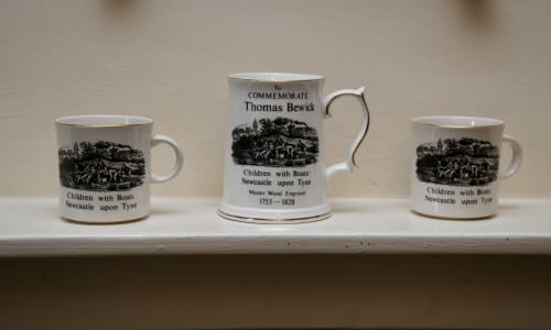 Three commemorative porcelain mugs featuring Thomas Bewick's 'Children with Boats' Newcastle upon Tyne engraving displayed on