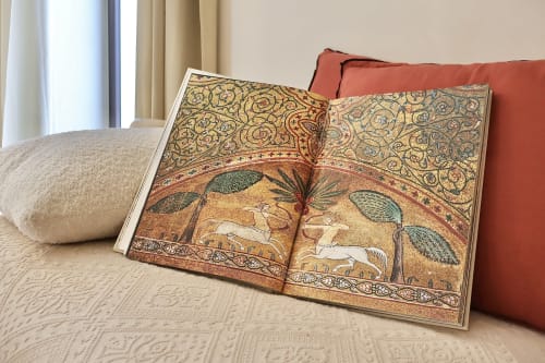 Libro riccamente decorato aperto su biancheria color panna con cuscini rossi nella Suite Deluxe Tagasuites, Catania