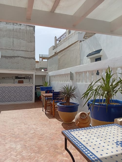 La cour ensoleillée du Riad Le Village Mogador avec ses jardinières bleues, table en mosaïque et balustrades blanches à Essaouira