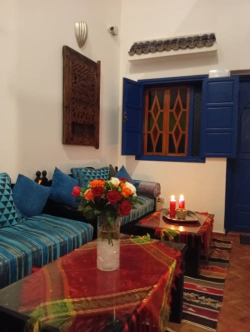 Salon du Riad Le Village Mogador avec fenêtre aux volets bleus, canapé turquoise rayé, table drapée rouge et fleurs fraîches
