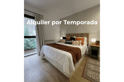 Dormitorio moderno en Vielha con cama queen, cabecero de madera, paredes beige, ventana grande con cortinas, mesita de noche y piso de madera