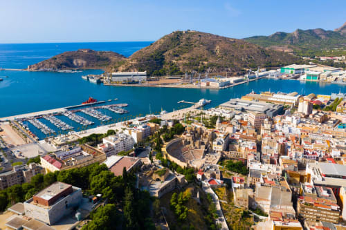 Vista aérea del puerto histórico de Cartagena con marina, anfiteatro y costa mediterránea rodeada de colinas.