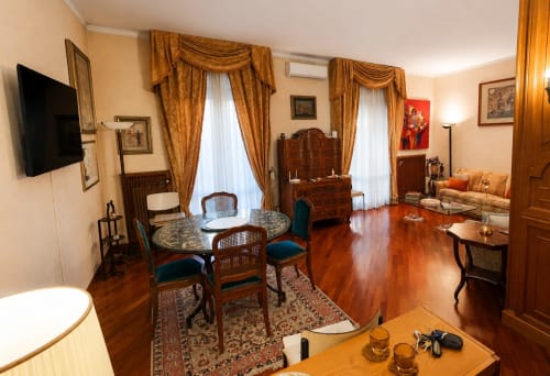 Elegant living room in House Mary Grace apartment, Roma. Hardwood floors, gold drapes, wooden furniture, TV, and garden view.