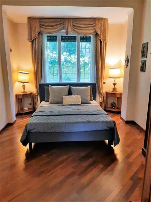 Elegante camera da letto in appartamento romano con vista giardino, pavimenti in legno, letto nero, illuminazione calda e finestre con drappeggi