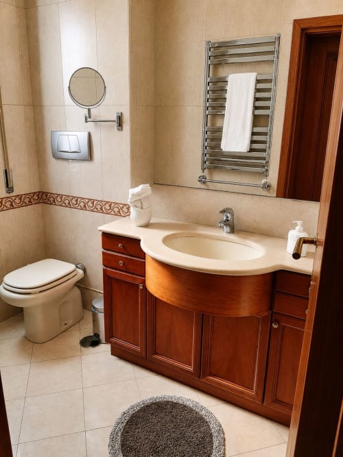 Modern bathroom in Rome apartment with curved wooden vanity, heated towel rack, circular mirror, and cream tile flooring