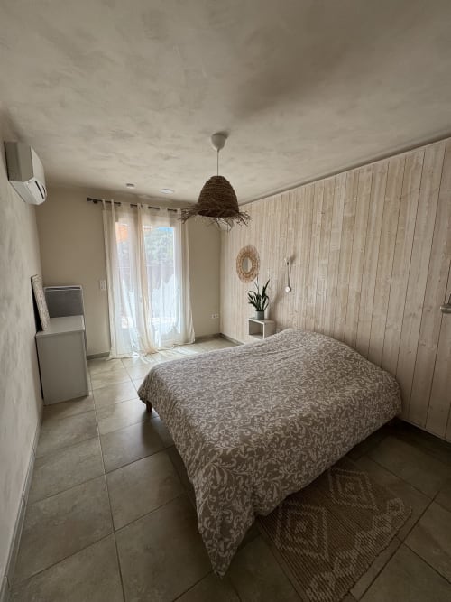 Chambre lumineuse avec parure de lit à motifs, murs boisés, luminaire suspendu, climatisation et vue sur jardin au Domaine d'Olga