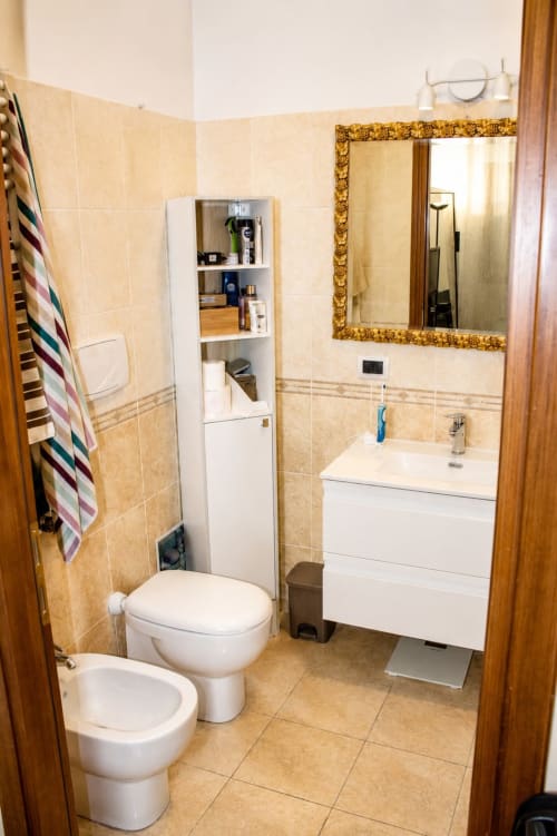 Modern bathroom in Rome apartment with white vanity, toilet, gold-framed mirror, storage shelves, and beige tile