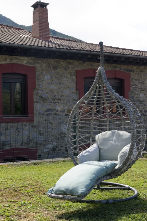 Silla colgante tejida gris con cojines en el jardín de Casa Rural Posada Real de Pajares, con vistas a las montañas y edificio de piedra