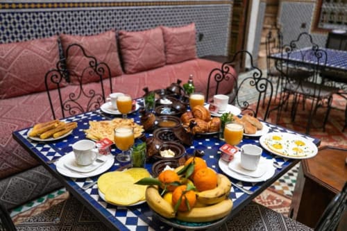 Table du petit-déjeuner au Riad Fes Aicha avec viennoiseries, fruits frais, jus et œufs surplombant des sièges aux coussins roses à Fès
