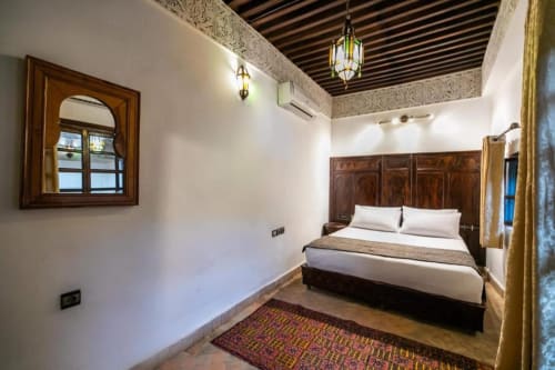 Chambre du Riad Fes Aicha avec lit en bois sculpté, murs blancs, plafond décoratif, lanterne suspendue et tapis marocain traditionnel
