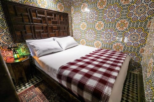Chambre du Riad Fes Aicha à Fès avec murs en zellige orné, tête de lit en bois sculpté, literie écossaise bordeaux