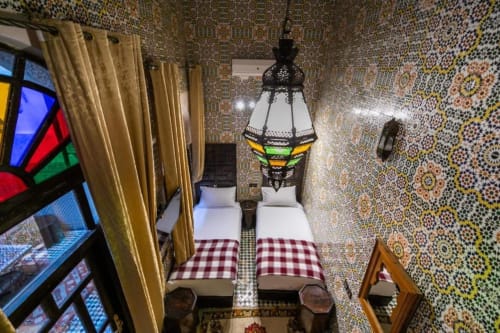 Chambre du Riad Fes Aicha avec lits jumeaux, zellige marocain orné, rideaux dorés et lanterne noire décorée.