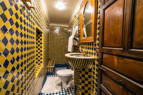 Salle de bains aux carreaux en losange bleu et jaune du Riad Fes Aicha à Fès, Maroc, avec une porte en bois ouvragée