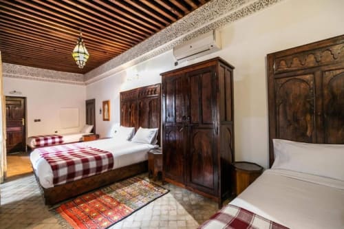 Chambre du Riad Fès Aïcha avec lits en bois sculpté traditionnel, literie à carreaux rouges et plafond et zellige marocain