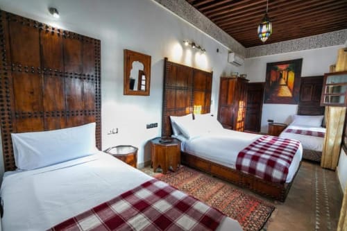 Chambre twin du Riad Fes Aicha avec portes en bois ouvragé, lits blancs, couettes bordeaux et décor marocain à Fès.