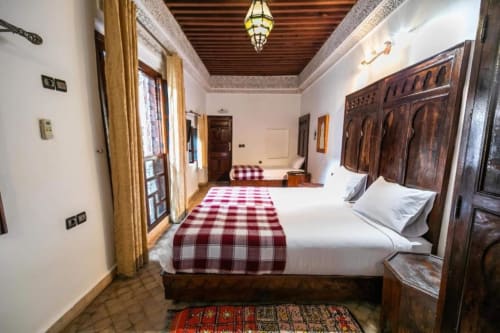 Chambre d'hôtel Riad Fes Aicha avec lits jumeaux, chemin de lit à carreaux rouges, portes en bois sculptées et luminaire marocain ouvragé.