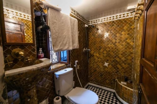 Salle de bain du Riad Fes Aicha avec carrelage mosaïque doré, toilettes blanches, lavabo et sol à damier noir