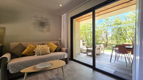 Studio moderne au Domaine Lantana avec canapé gris, coussins jaunes et portes coulissantes ouvertes sur la terrasse du jardin