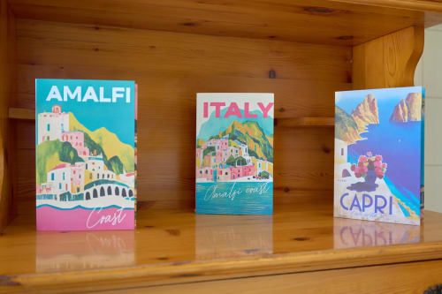 Three colorful travel posters of Italian coastal destinations displayed on wooden shelf in The Lemon Garden Escape villa at