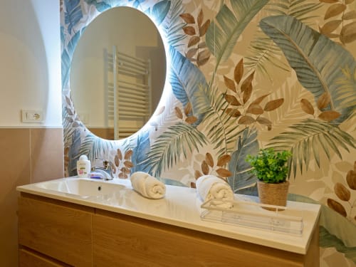 Modern bathroom vanity with illuminated oval mirror, tropical leaf wallpaper, rolled towels, and potted plant at Sorrento Sea