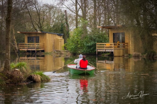 Deux clients font du canotage dans un bateau vert entre des cabanes en bois sur l'eau aux Rives du bel air, un gîte fluvial français