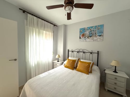 Dormitorio luminoso con ropa de cama blanca, almohadas doradas, ventilador de techo y arte costero en apartamento de Fuengirola
